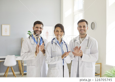 Three smiling doctors in white coats standing in medical office, clapping hands, celebrating success Three smiling doctors in white coats standing in medical office, clapping hands, celebrating success 128705745