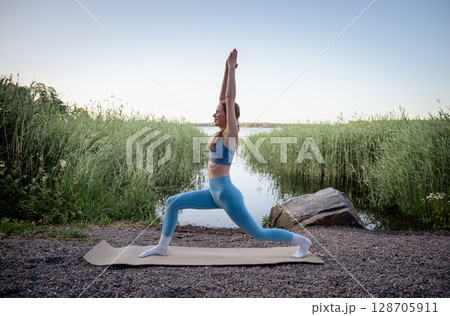 Young woman doing yoga exercise outdoors near lake in summer  128705911