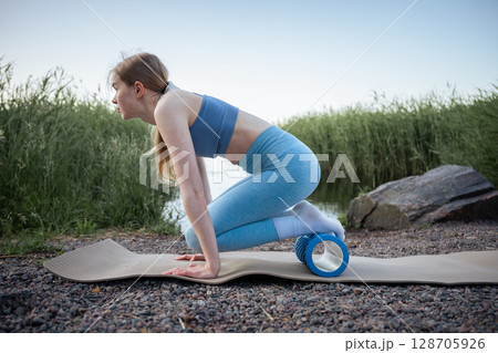Woman using foam massage roller on yoga mat by summer lake 128705926
