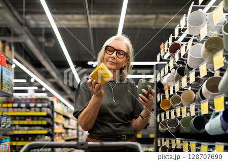 Stylish mature woman shopping for home goods in a modern retail store selects decor and kitchen Stylish mature woman shopping for home goods in a modern retail store selects decor and kitchen 128707690