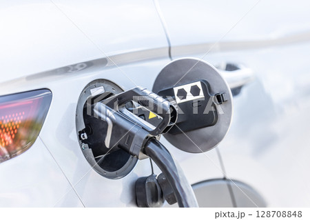 Focus closeup electric vehicle plugged in with EV charger device from blurred background of public charging station powered by renewable clean energy for progressive eco-friendly car concept 128708848