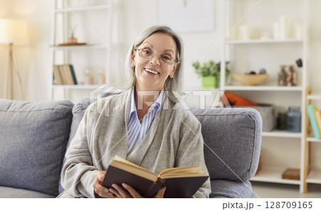 Happy senior woman sitting on sofa at home and reading a book in living room 128709165