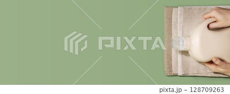Eco friendly washing banner. Female hands hold bottle with washing gel or fabric softener on a textile napkins on a green background with copy space. Laundry day. Flat lay, banner size Eco friendly washing banner. Female hands hold bottle with washing gel or fabric softener on a textile napkins on a green background with copy space. Laundry day. Flat lay, banner size 128709263