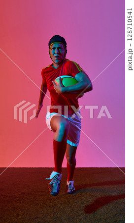 Female rugby player running with ball under vibrant pink and purple lighting 128710541
