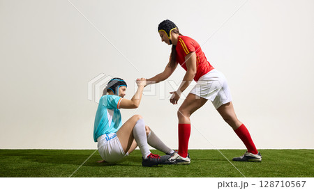 Female rugby athlete helping another to get up on grassy field in match Female rugby athlete helping another to get up on grassy field in match 128710567