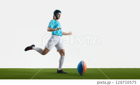 Female rugby player striking a ball on grassy field with dynamic movement 128710575