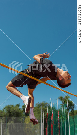 Athlete executing high jump beneath clear blue sky at outdoor track area 128710588