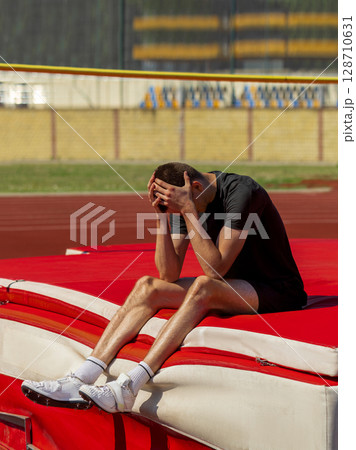 Exhausted athlete sitting on high jump mat during track and field practice Exhausted athlete sitting on high jump mat during track and field practice 128710631