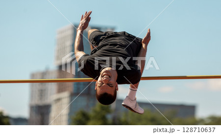 Athlete executes high jump with urban scenery on sunny day 128710637