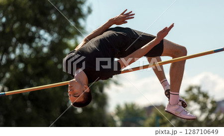 Athlete in mid-air clearing high jump bar under clear sky 128710649