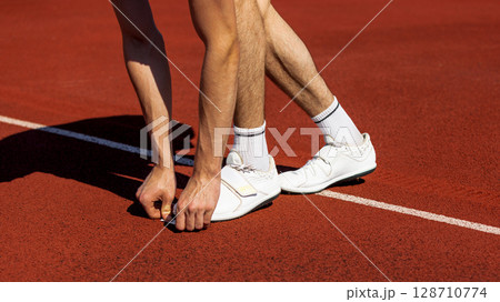 Athlete in starting stance on red track surface under bright sunlight preparing to sprint 128710774