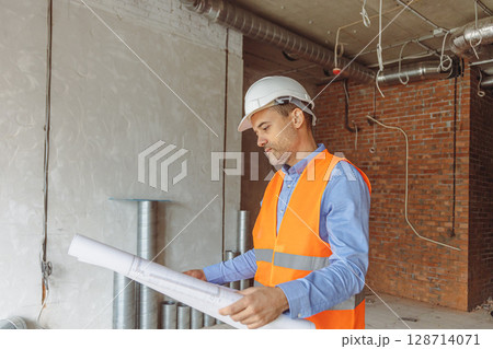 Construction worker reviewing blueprints in a partially completed building in a busy urban setting Construction worker reviewing blueprints in a partially completed building in a busy urban setting 128714071