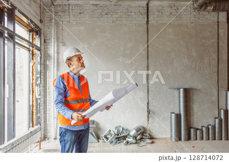 Construction worker analyzes blueprints in a newly built office space during daylight hours 128714072