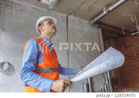Construction manager reviewing plans in a partially finished building with exposed walls and pipes 128714075
