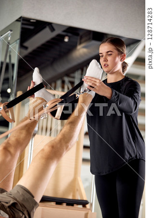 Professional fitness instructor assisting athlete during pilates workout using reformer equipment 128714283