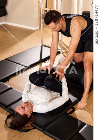 Trainer assisting with fitness exercise on a mat in a gym setting 128714477