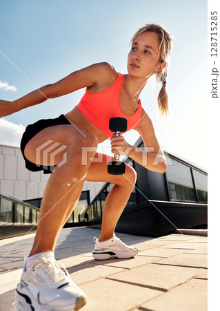 Determined athletic woman training outside with weights on a sunny day Determined athletic woman training outside with weights on a sunny day 128715285