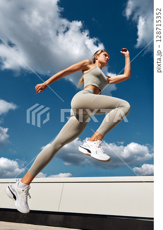 Woman in sportswear leaping outside with a blue sky and clouds background 128715352