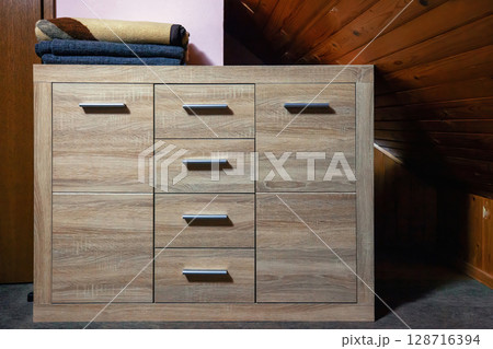 Wooden chest of drawers with folded blankets on top in small attic bedroom, front view 128716394