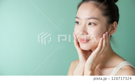 Woman Touching Face with a Soft Smile on a Light Green Background in Natural Light Setting 128717324