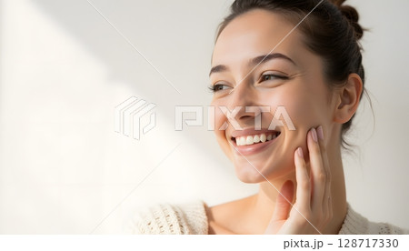 Happy young woman smiling gently while touching her face in natural light indoors 128717330
