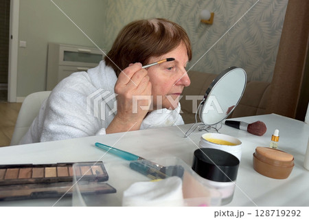 An older woman with charm is sitting in her living room, doing her makeup routine. An older woman with charm is sitting in her living room, doing her makeup routine. 128719292