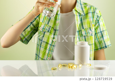 Organic kids vitamins and nutrients. Mockup of natural bio pills, vitamins or supplements. The boy washes down vitamins in gel capsules with water. Supplements of Omega 3 or D 3 Organic kids vitamins and nutrients. Mockup of natural bio pills, vitamins or supplements. The boy washes down vitamins in gel capsules with water. Supplements of Omega 3 or D 3 128719519