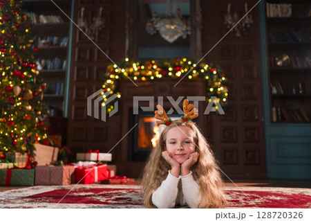 Merry Christmas. Little girl smiling near Christmas tree in classical dark interior. Young happy kid wearing deer horns in living room with fireplace Christmas tree gift boxes. Christmas eve at home Merry Christmas. Little girl smiling near Christmas tree in classical dark interior. Young happy kid wearing deer horns in living room with fireplace Christmas tree gift boxes. Christmas eve at home 128720326