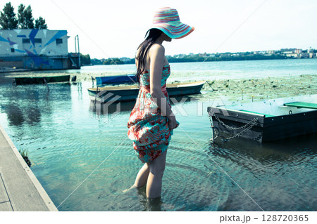 A young girl in a long colorful dress and a hat stands barefoot in the clear water of the lake A young girl in a long colorful dress and a hat stands barefoot in the clear water of the lake 128720365
