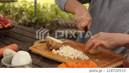 Cutting fresh onion for preparation of vegetable dish in outdoor kitchen setting surrounded by nature showcasing skillful hands in action Cutting fresh onion for preparation of vegetable dish in outdoor kitchen setting surrounded by nature showcasing skillful hands in action 128720513