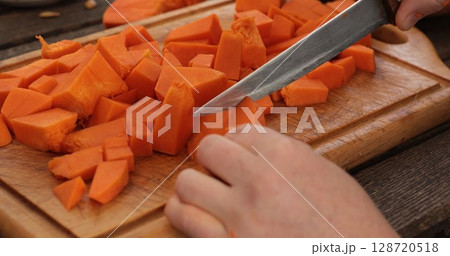 Chopping bright orange pumpkin pieces on wooden cutting board with sharp knife reflecting effort and precision, early autumn kitchen preparations create warm atmosphere for seasonal dishes Chopping bright orange pumpkin pieces on wooden cutting board with sharp knife reflecting effort and precision, early autumn kitchen preparations create warm atmosphere for seasonal dishes 128720518