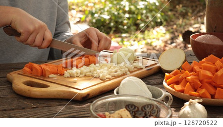 Chopping vegetables outdoors during sunny afternoon, focusing on preparation for healthy meal, featuring carrots, onions, squash, and fresh herbs on wooden cutting board 128720522