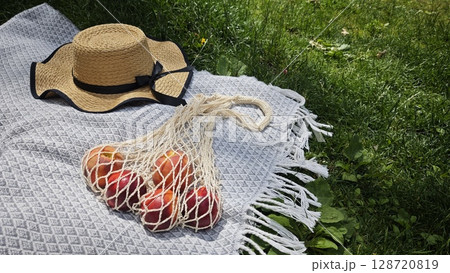 Straw hat with black ribbon and string bag of nectarines on patterned blanket on grass 128720819