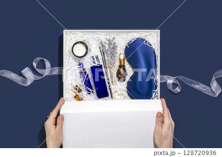 Men holiday gift box. A female hands hold a white gift box with sleep care set for men. Preparing self care package, craft gift box with mask for sleep, aroma candle, aroma sticks, oil and bubble bath 128720936