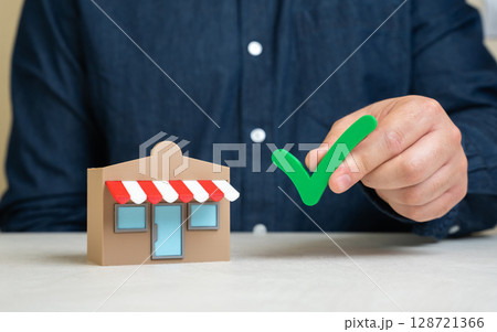 A man holds a green check mark near a store. Pass the test. Quality verification. Business permits. Successful inspection, business licensing, or compliance with regulations. Audit approval A man holds a green check mark near a store. Pass the test. Quality verification. Business permits. Successful inspection, business licensing, or compliance with regulations. Audit approval 128721366