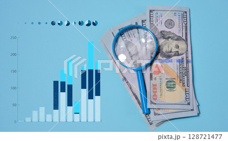 A stack of hundred dollar bills lies on a blue background, a magnifying glass, next to it is a vertical histogram with graphs. A stack of hundred dollar bills lies on a blue background, a magnifying glass, next to it is a vertical histogram with graphs. 128721477