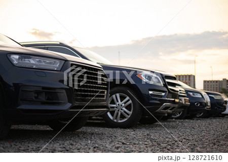 A row of dark-colored SUVs parked outside on a gravel lot. The cars are lined up, presenting a view of their front ends. 128721610