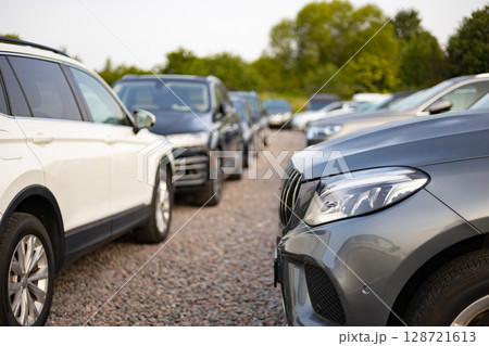 A row of various cars for sale outdoors, potentially at a car dealership. The vehicles are parked on a gravel surface with trees in the background. A row of various cars for sale outdoors, potentially at a car dealership. The vehicles are parked on a gravel surface with trees in the background. 128721613