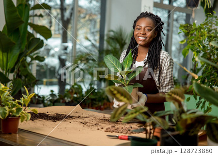 Cute african american woman in apron standing at the desk with plants Cute african american woman in apron standing at the desk with plants 128723880