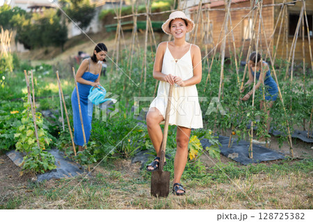 Girl near bed of greenhouse plants. Two garden plot workers are busy in background 128725382