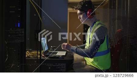 Operating laptop, technician wearing neon vest red earmuffs in server room with racks, copy space 128729666