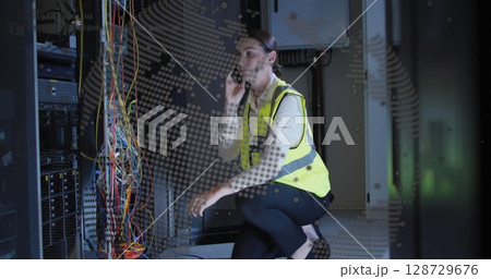 Kneeling female tech wearing yellow vest routing network cables in data center, with smartphone 128729676