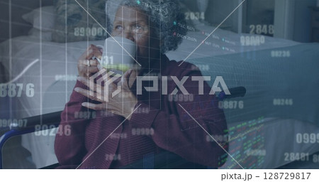 Holding ceramic mug senior African American woman in wheelchair gazing in lit bedroom, data overlay 128729817