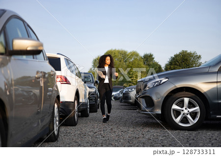 A businesswoman walks through a car lot, possibly selling vehicles. She is looking at the cars, possibly for a customer. A businesswoman walks through a car lot, possibly selling vehicles. She is looking at the cars, possibly for a customer. 128733311
