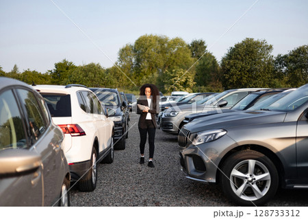 A businesswoman reviews paperwork between rows of cars in a dealership lot, assessing vehicle options. 128733312