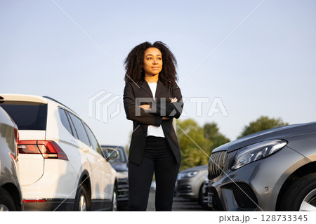 A confident woman in a business suit standing near cars, possibly a car saleswoman or manager, with a look of assurance. A confident woman in a business suit standing near cars, possibly a car saleswoman or manager, with a look of assurance. 128733345