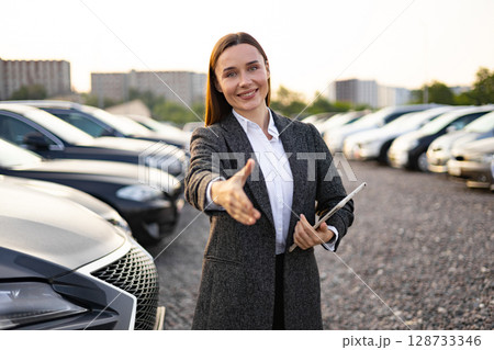 A businesswoman offers a handshake at a car dealership, symbolizing a successful car sale or purchase. A businesswoman offers a handshake at a car dealership, symbolizing a successful car sale or purchase. 128733346