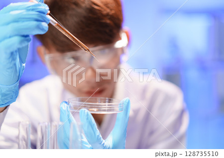 Female lab technician holding pipette above Petri dish. Young scientist doing analysis in the laboratory. Female lab technician holding pipette above Petri dish. Young scientist doing analysis in the laboratory. 128735510