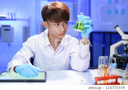 A young male scientist examining a in beaker a lab. A young male scientist examining a in beaker a lab. 128735554