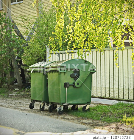 green trash containers in sunlit street near fence with birch tree branches and greenery green trash containers in sunlit street near fence with birch tree branches and greenery 128735728